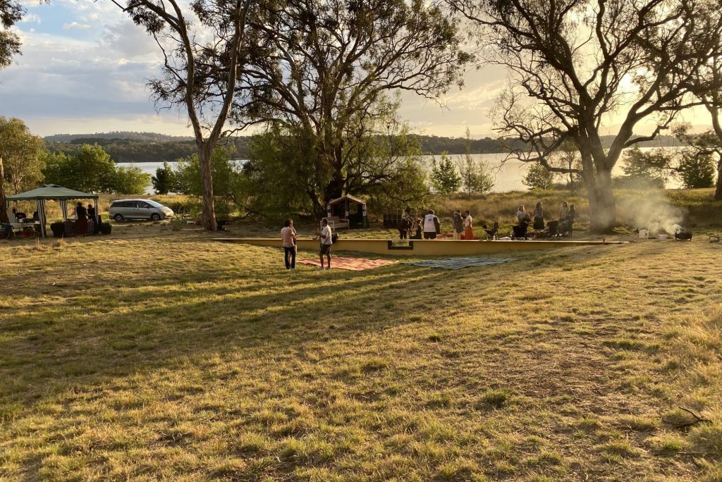 It is outside, evening on a summer day on Ngunnawal Country. The sky buttered with a few clouds but it is clearly warm, the evening sun yellowing everything through the cloud cover. Big ngunnawal gumtrees lean over a scene of people in the middle-distance, sitting on mats, standing around and being together. You can learn more about the scene in the article.