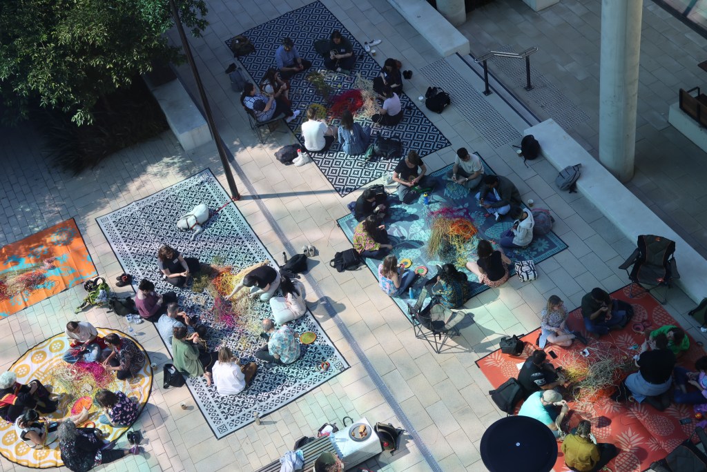 A birds-eye view of people sitting in circles outdoors, in a paved area, weaving all across the area. Gaanyatjarra lore women Dr Elizabeth Marrkilyi Ellis and Aunty Nerida Giles leading knowledge making and sharing sessions at AusSTS 2024.